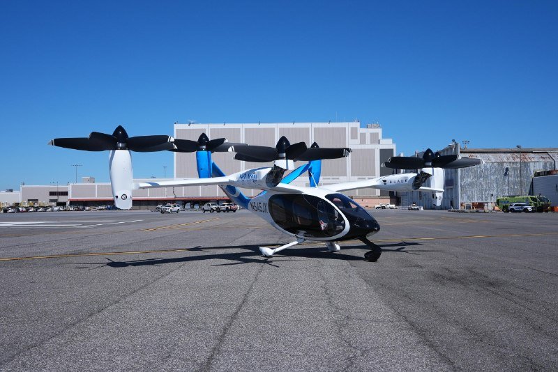 A white and blue electric air taxi aircraft with multiple rotors sits on a tarmac in front of a large hangar under a clear blue sky.