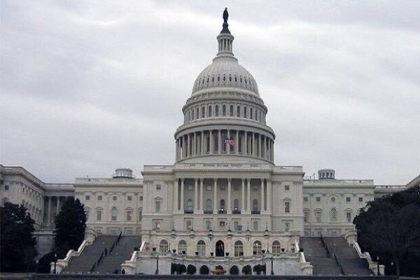 The United States Capitol building stands beneath an overcast sky, its white neoclassical dome and columns prominently visible from the front steps.