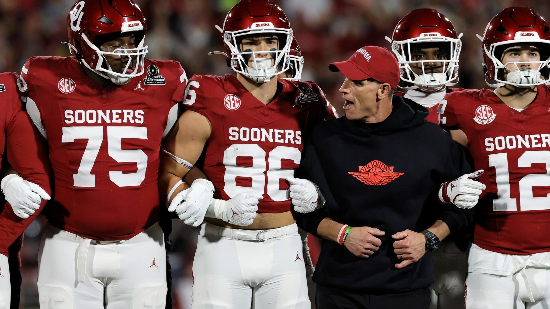 Oklahoma Sooners football players in crimson jerseys stand with a coach in a black Air Jordan hoodie during a college football game.