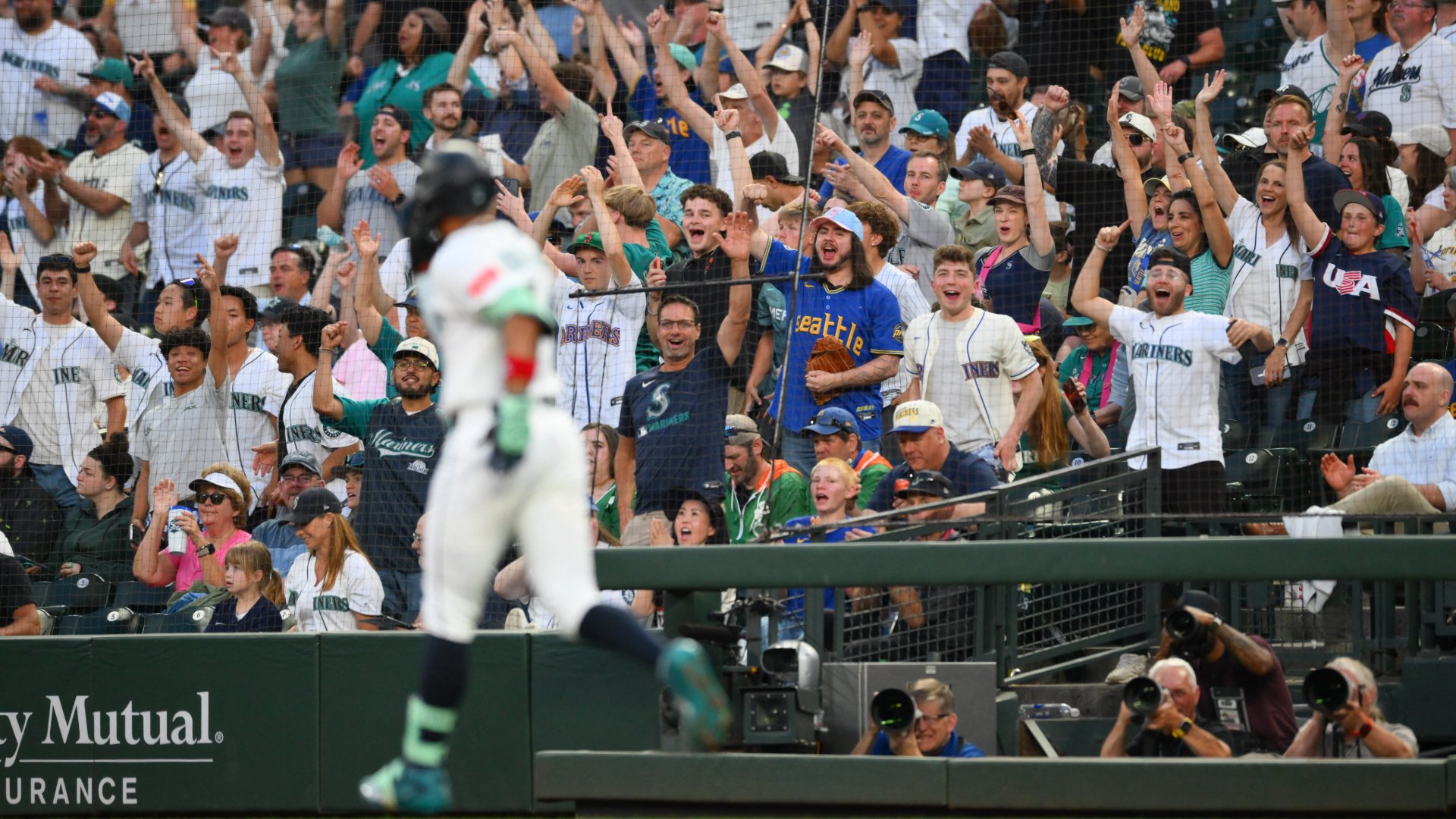 A baseball player in a white uniform runs past a cheering crowd wearing Mariners jerseys with arms raised behind a protective net at a stadium.