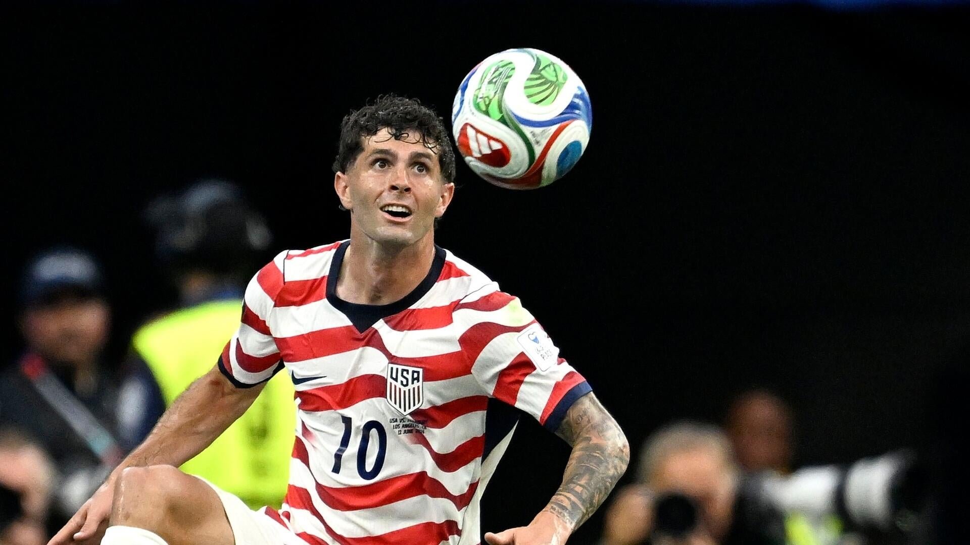 A USA soccer player wearing jersey number 20 dribbles the ball past two opposing defenders in dark blue kits during a match in front of a stadium crowd.