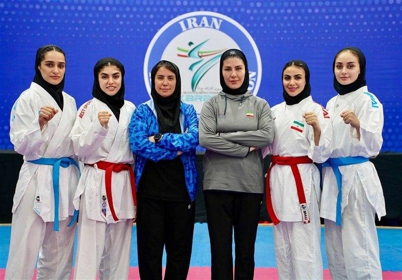 Six women in martial arts uniforms and hijabs pose together indoors, with two in tracksuits in the center and four in white gi uniforms with blue and red belts, before a blue backdrop featuring the word "IRAN."