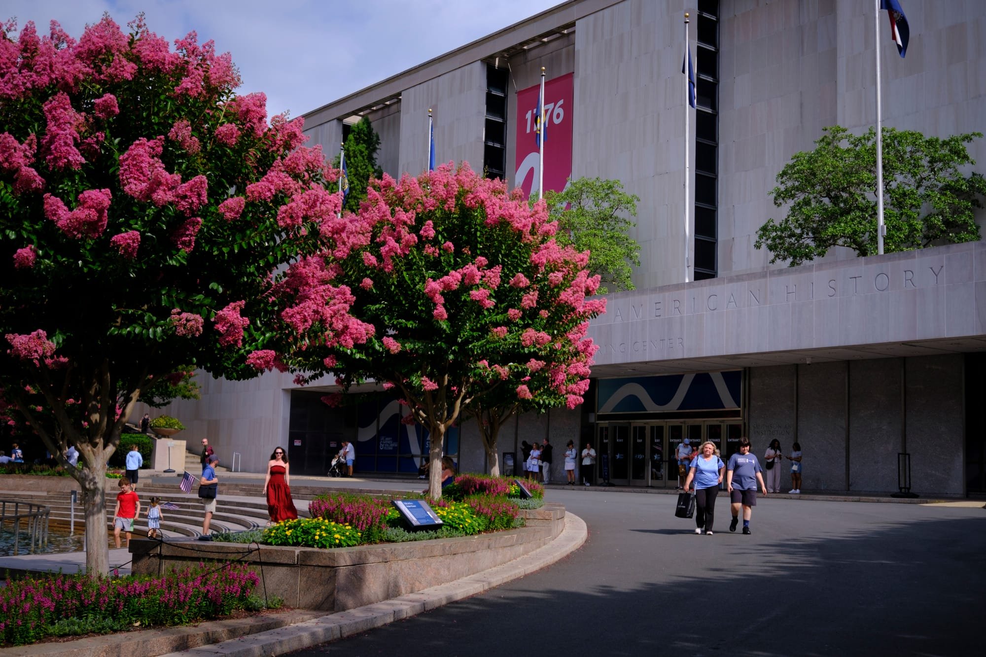 Visitors walk past pink-flowering crepe myrtle trees outside the stone entrance of the Smithsonian National Museum of American History building, with a "1776" banner displayed above.