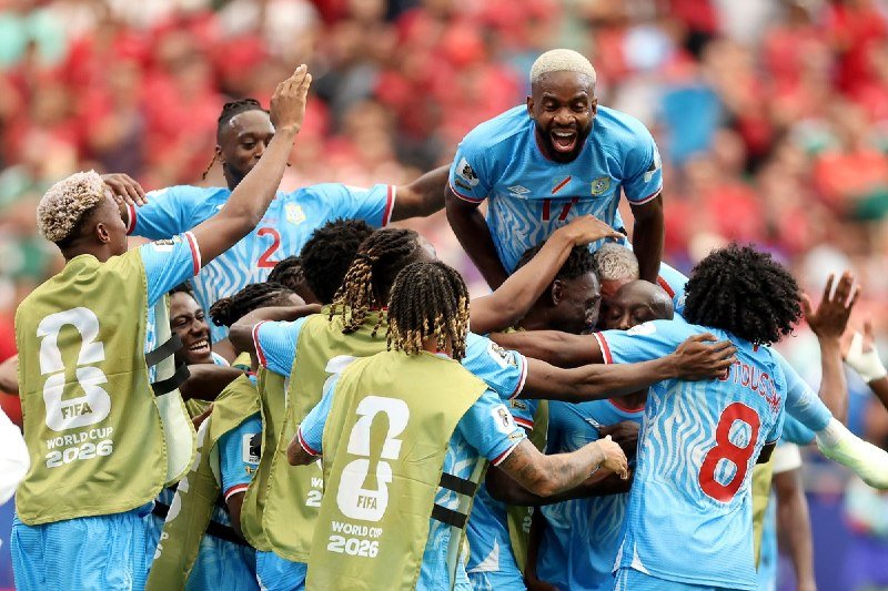 Four soccer players in white jerseys with red and green trim embrace and celebrate joyfully in a stadium, with one wearing a gold "FIFA World Cup 2025" bib.