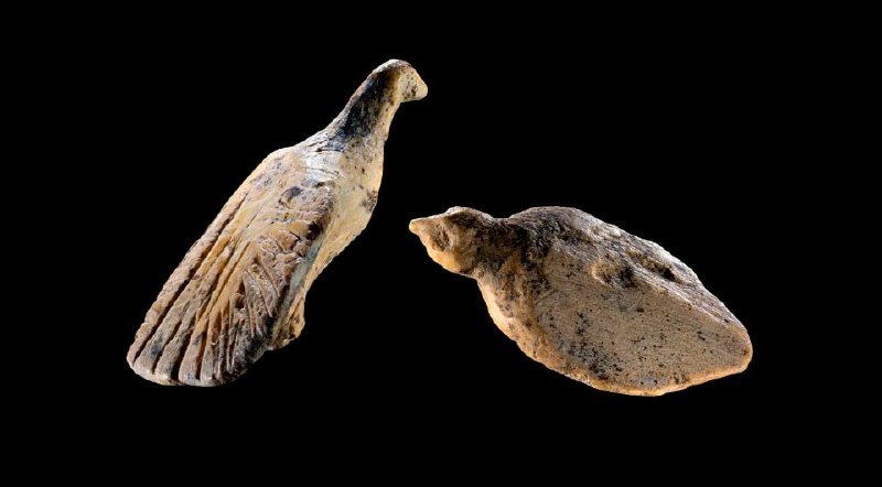 Two weathered stone or clay bird-shaped artifacts, one with carved wings and a crest, rest against a black background.