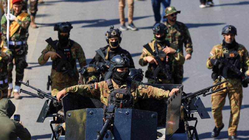 Armed soldiers in camouflage uniforms, helmets, and face coverings stand on a paved street, some holding rifles and positioned behind a mounted machine gun.