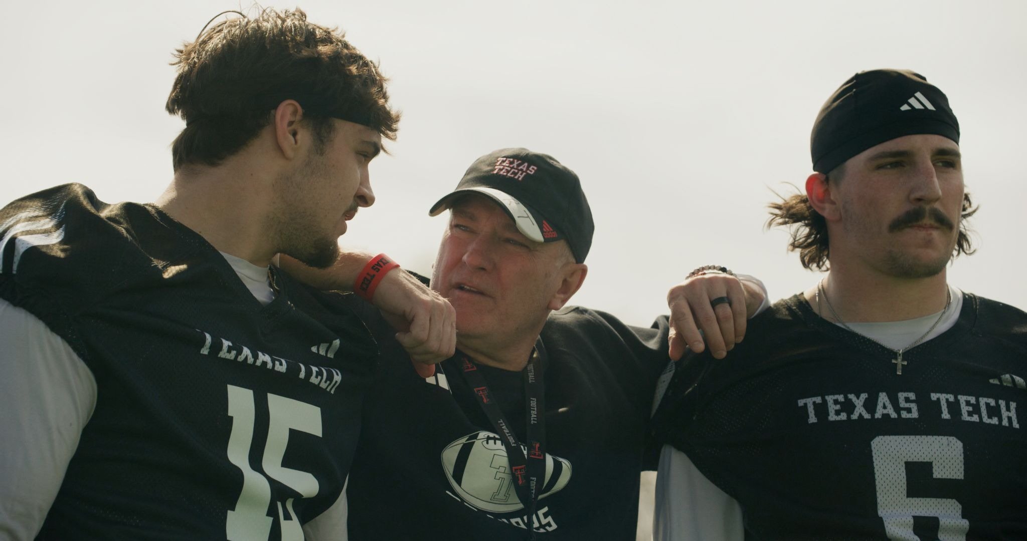 A man in a "Texas Tech" cap stands between two football players in black "Texas Tech" jerseys, his arms around their shoulders.