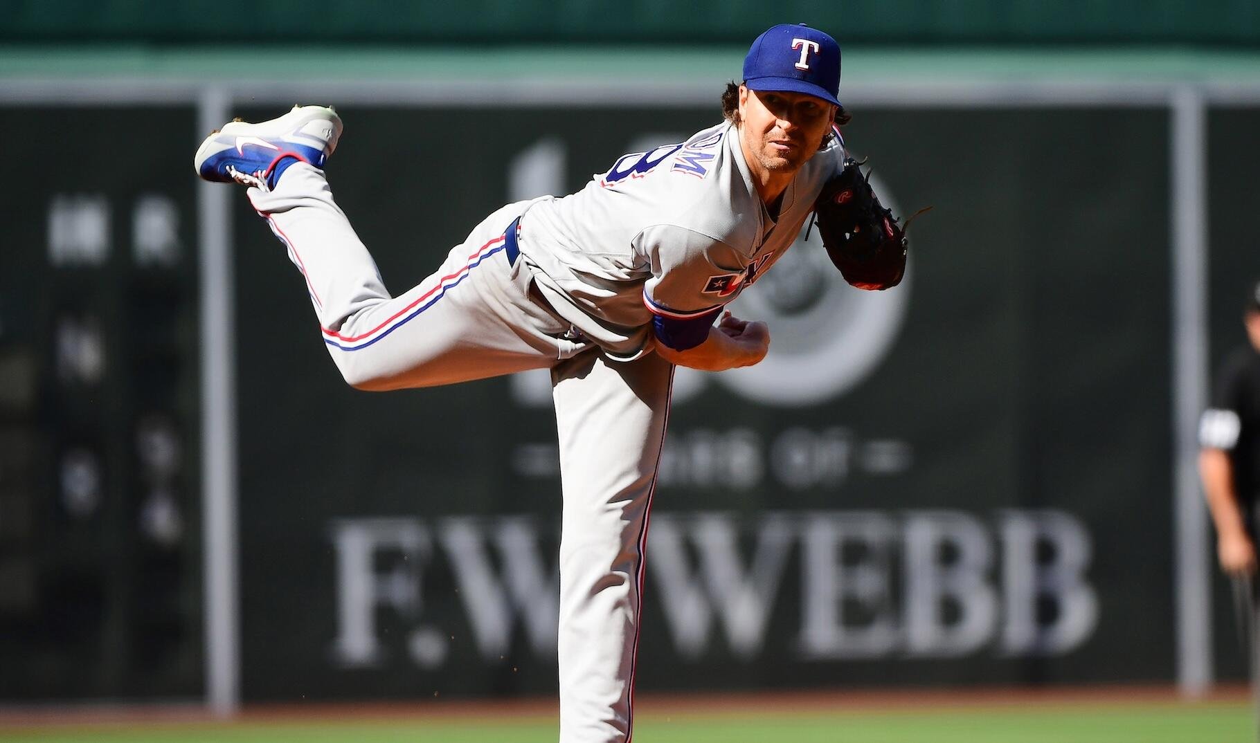 A Texas Rangers pitcher in a gray uniform follows through on a delivery at a baseball stadium, with an outfield wall visible behind him.
