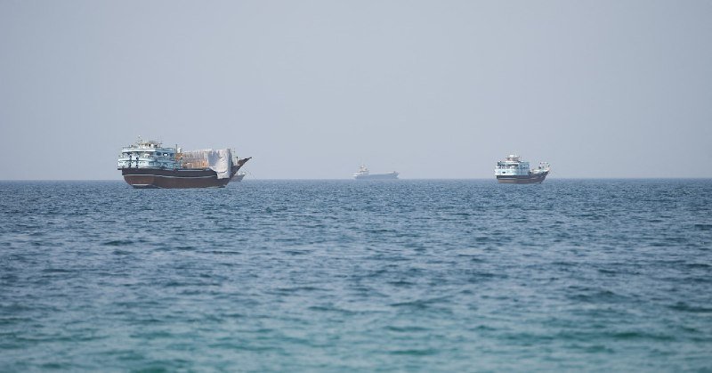 Three large cargo ships are anchored in a calm, hazy sea under an overcast sky.