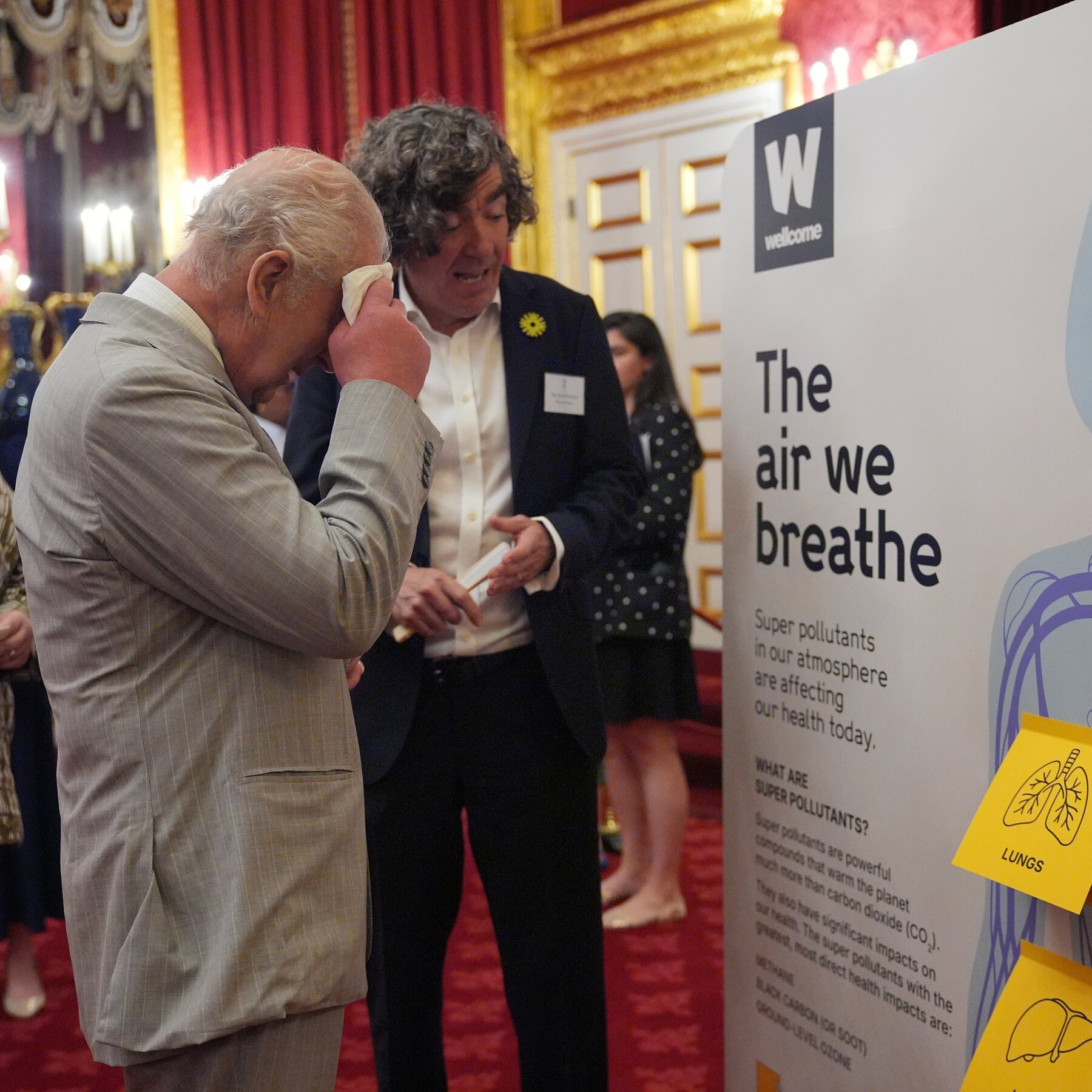 A man in a gray suit wipes his face with a tissue beside a Wellcome display titled "The air we breathe" in an ornate red-carpeted hall.