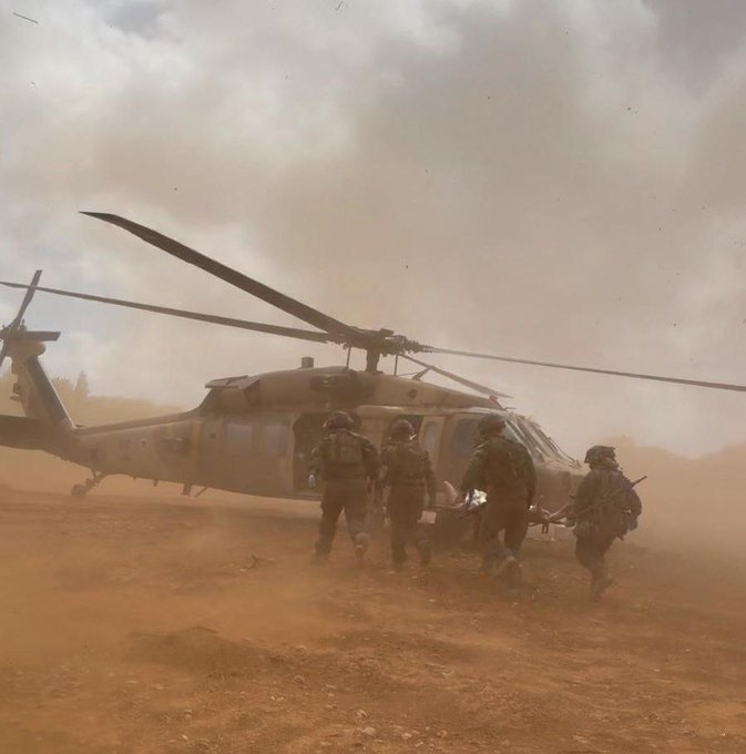 Soldiers in helmets and tactical gear walk toward a military helicopter amid thick dust and smoke in an outdoor setting.