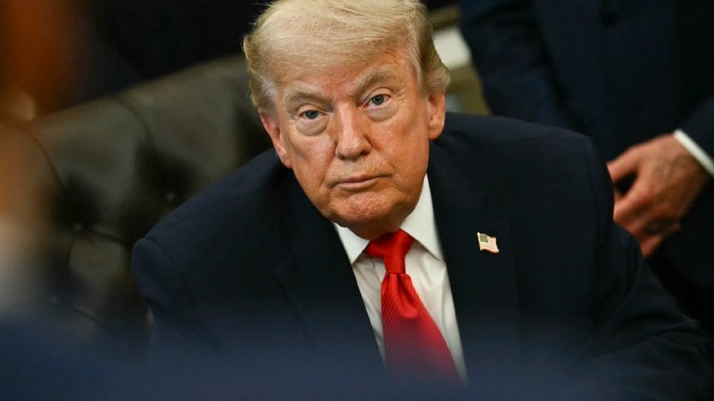A man with blond hair, wearing a dark suit, white shirt, and red tie with an American flag pin, sits indoors looking forward with a serious expression.