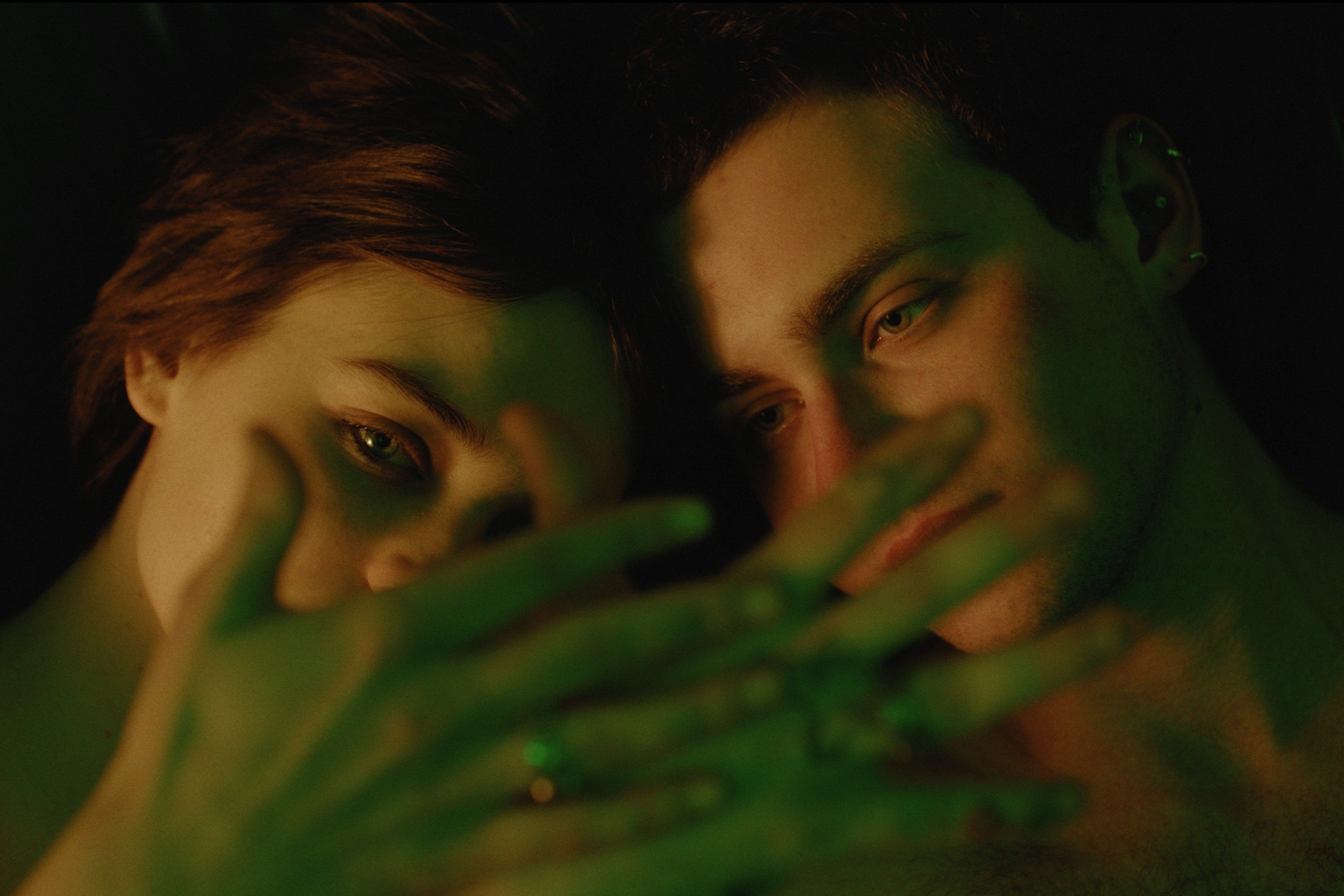 Two men are visible in close-up, their faces partially obscured by green leaves in the foreground under low lighting.