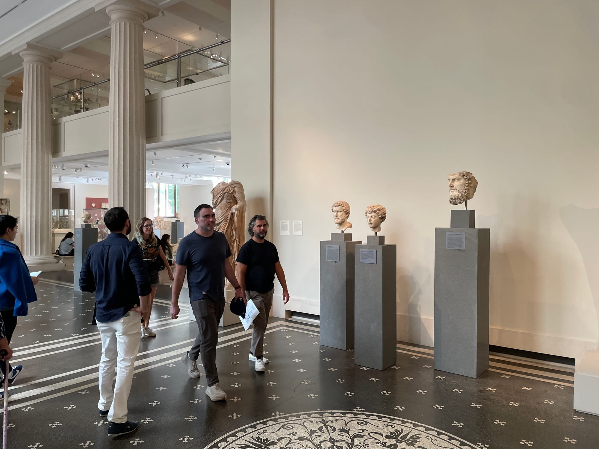 Visitors walk through a spacious museum gallery featuring tall classical columns, a patterned tile floor, and three ancient marble sculpted heads displayed on gray pedestals.