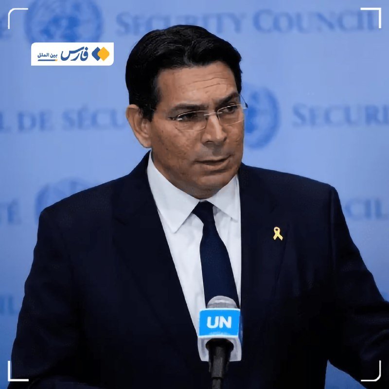 A man in a dark suit and glasses speaks into a UN-branded microphone, with a Security Council backdrop and a yellow ribbon pin on his lapel.