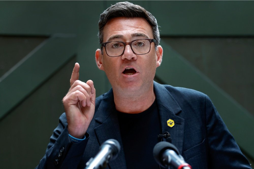 A bespectacled man in a dark blazer with a yellow pin gestures upward while speaking at microphones against a green geometric background.