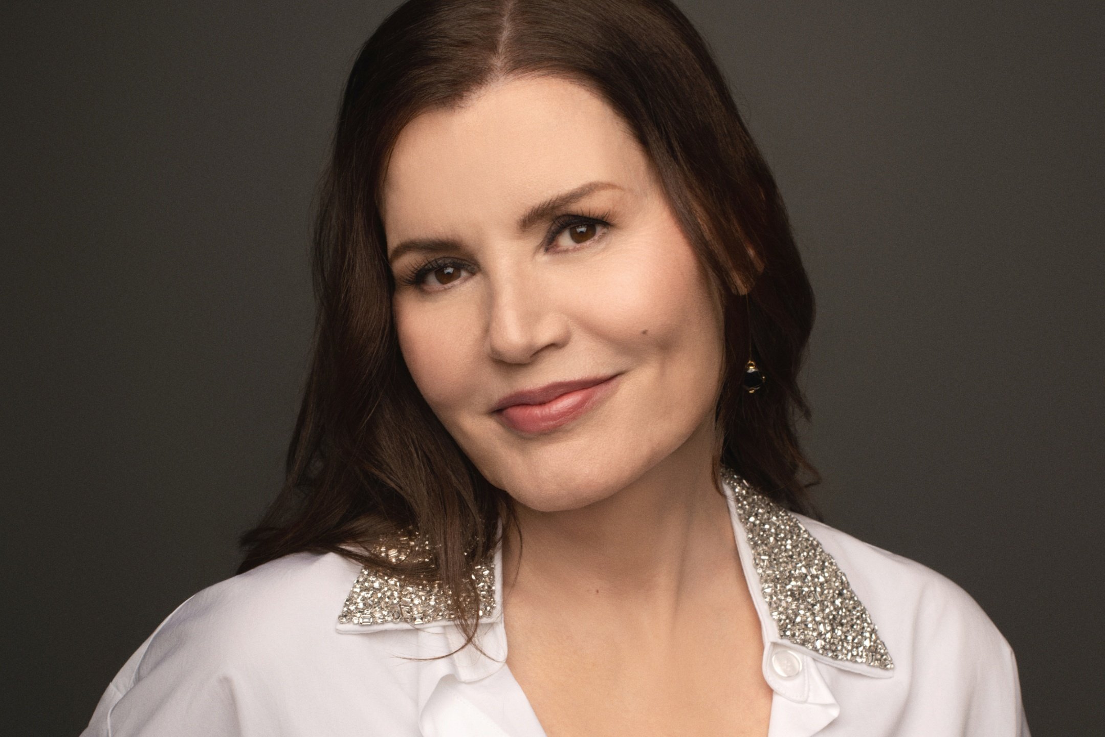 A woman with long brown hair poses against a gray background, wearing a white blouse featuring a rhinestone-embellished collar.