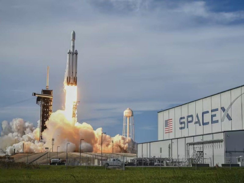 A SpaceX rocket lifts off from a launch pad, trailing bright flames and thick smoke beside a large hangar displaying the "SpaceX" logo and American flag.
