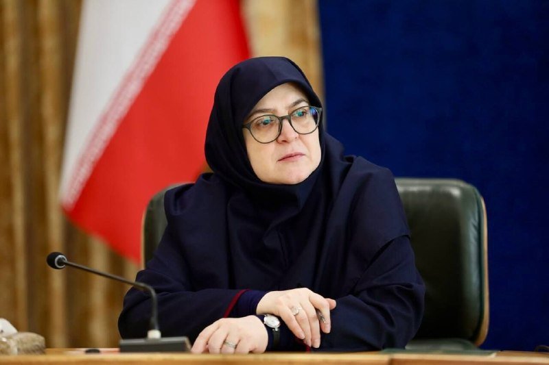 A woman wearing a dark hijab and glasses sits at a desk with a microphone, with a red and white flag visible behind her.