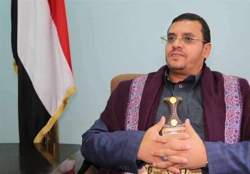 A bespectacled man in traditional patterned attire sits holding a wooden trophy against his chest, with a red, white, and black flag visible in the background.