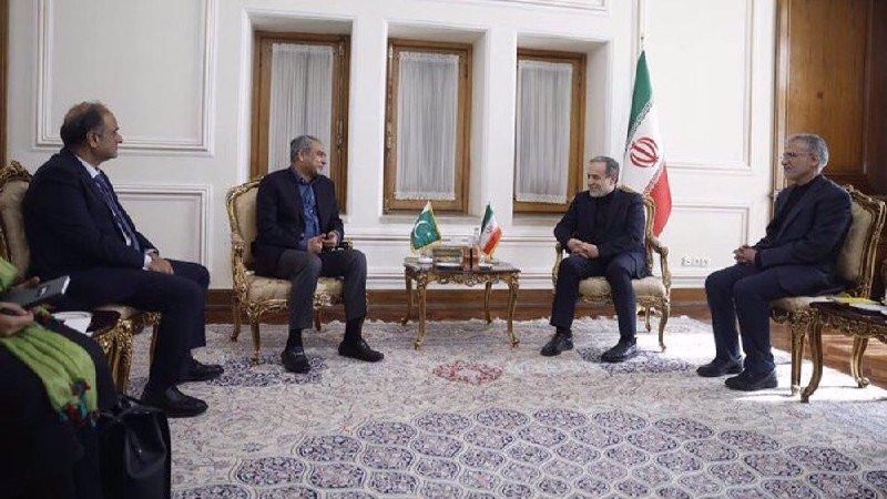 Four men in dark suits sit in ornate chairs in a formal room, facing each other with two flags displayed behind them.