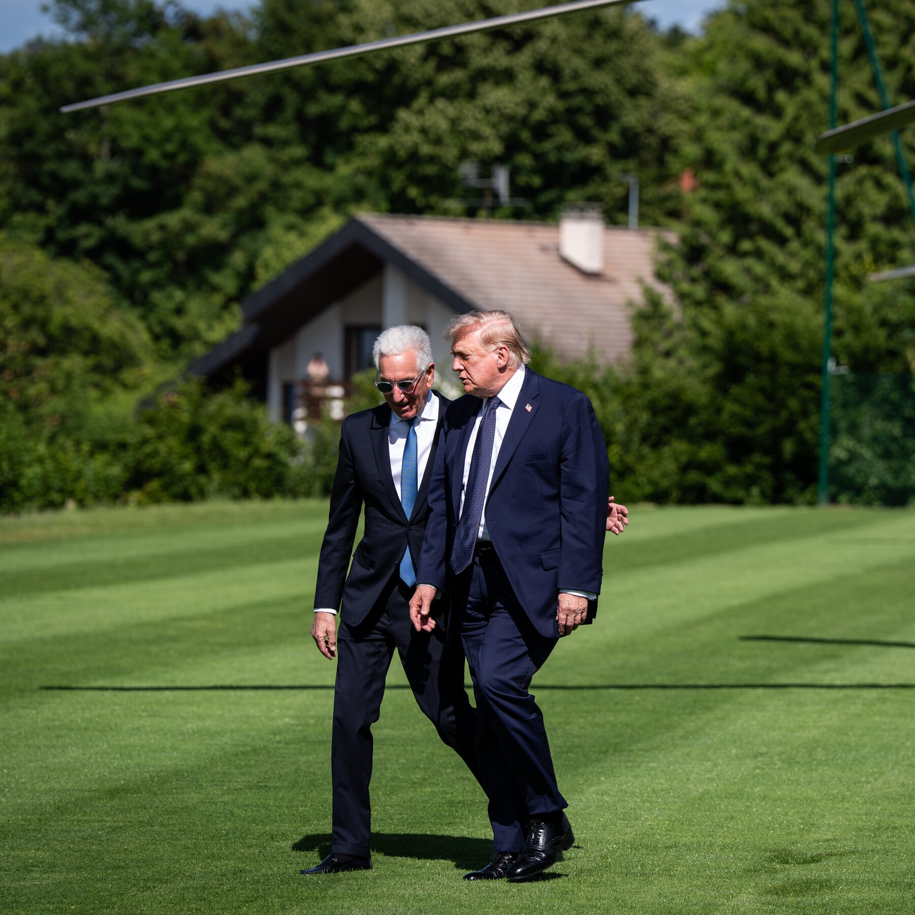 Two men in dark suits walk together across a green lawn, with a house and forested hillside in the background, one gesturing as they converse.