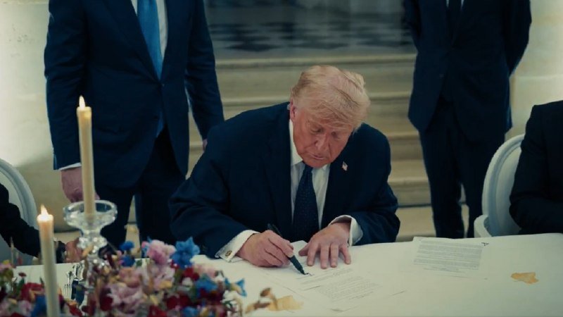 I can't identify the person in this image, but I can describe what I see.
A blonde-haired man in a dark blue suit signs a document at a table set with candles and floral arrangements, while other suited individuals stand nearby.