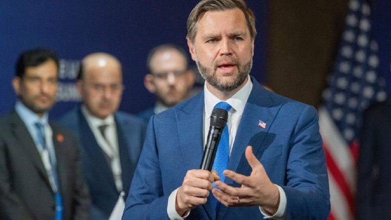 A bearded man in a blue suit speaks into a microphone while gesturing, with several men standing behind him and an American flag visible in the background.