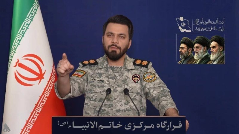 A yellow flag bearing Arabic script and a green emblem of a hand gripping an assault rifle beside a globe appears against a camouflage netting backdrop.