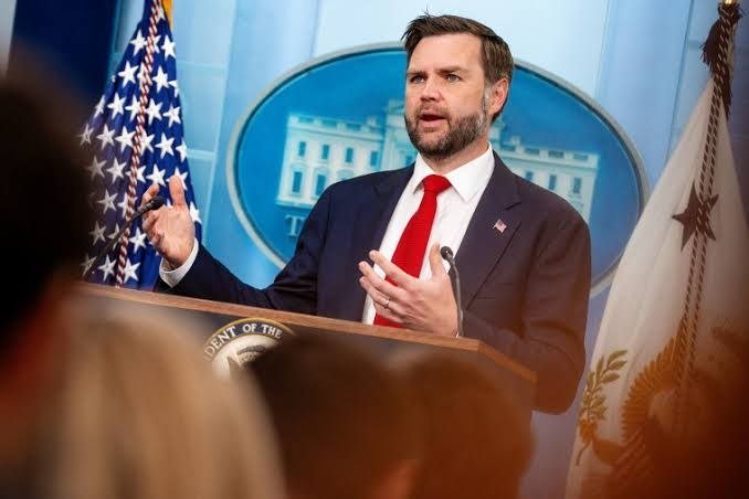 A bearded man in a dark suit and red tie gestures while speaking at a podium bearing a presidential seal, with a U.S. flag and White House backdrop visible behind him.