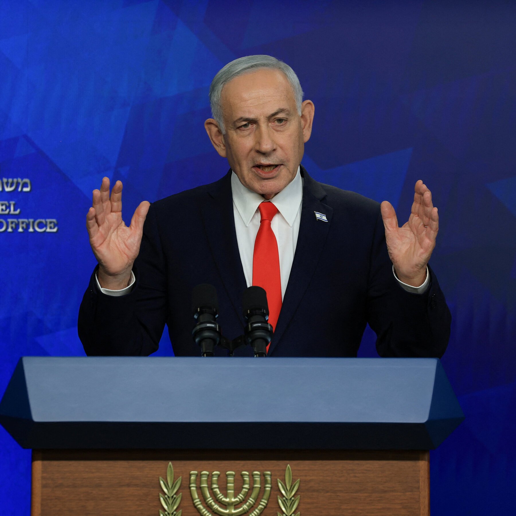 A gray-haired man in a dark suit and red tie gestures with both hands raised while speaking at a podium featuring a menorah emblem, set against a blue backdrop with partial "OFFICE" text.