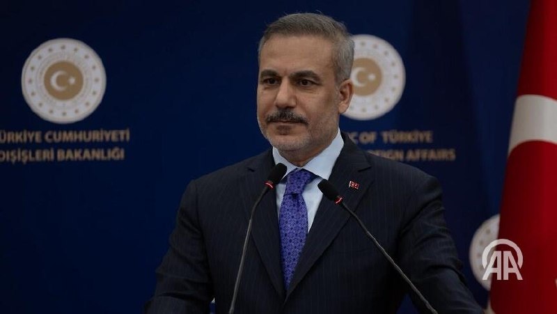 A gray-haired man in a dark suit and striped tie gestures while speaking at a conference table, with small national flags and other attendees visible around him.