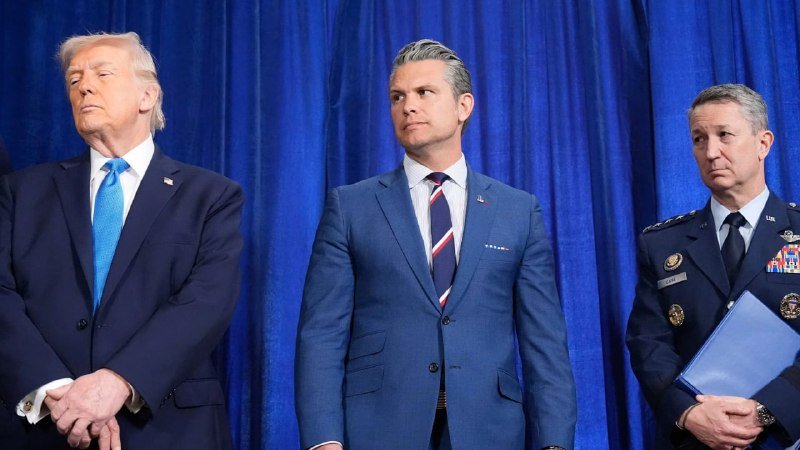 Three men—one in a dark suit with a blue tie, one in a blue suit with a striped tie, and one in a military uniform holding a folder—stand before a blue curtain.
