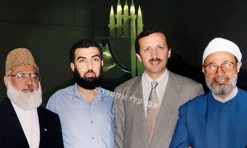 Four men pose together indoors, two wearing traditional Islamic caps and robes, two in Western suits, against a dark backdrop with Hebrew lettering.