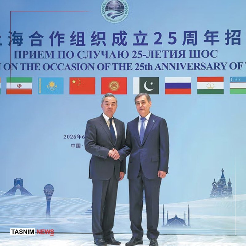 Two men in dark suits stand outdoors on a sunny day, with a person holding a camera and a crowd visible in the background.