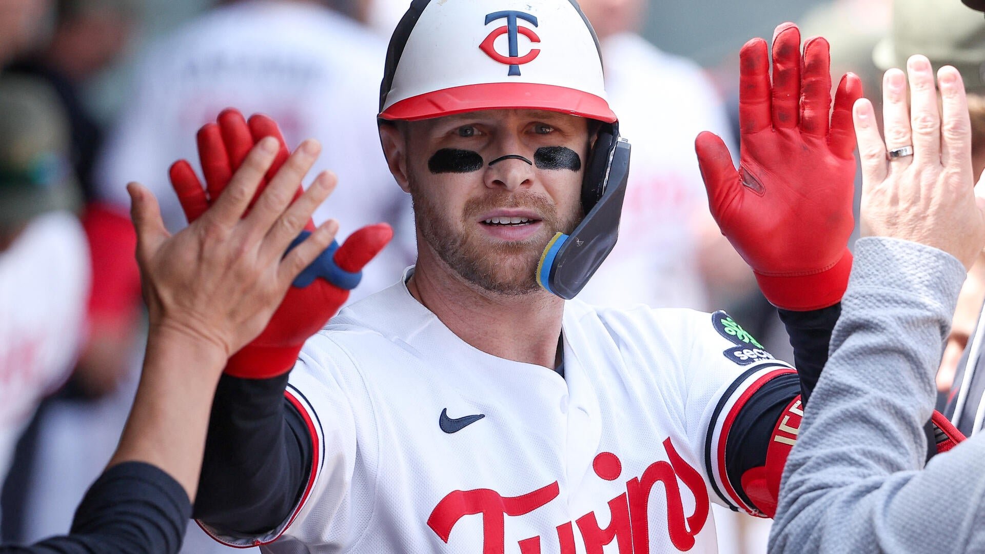 A Minnesota Twins baseball player in a white uniform and cap, wearing eye black and a protective face flap, raises his red-gloved hands to exchange high-fives in a dugout.