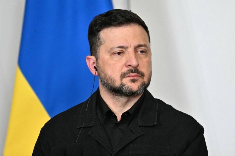 A dark-haired, bearded man in a black jacket wears an earpiece while standing before a blue and yellow flag.