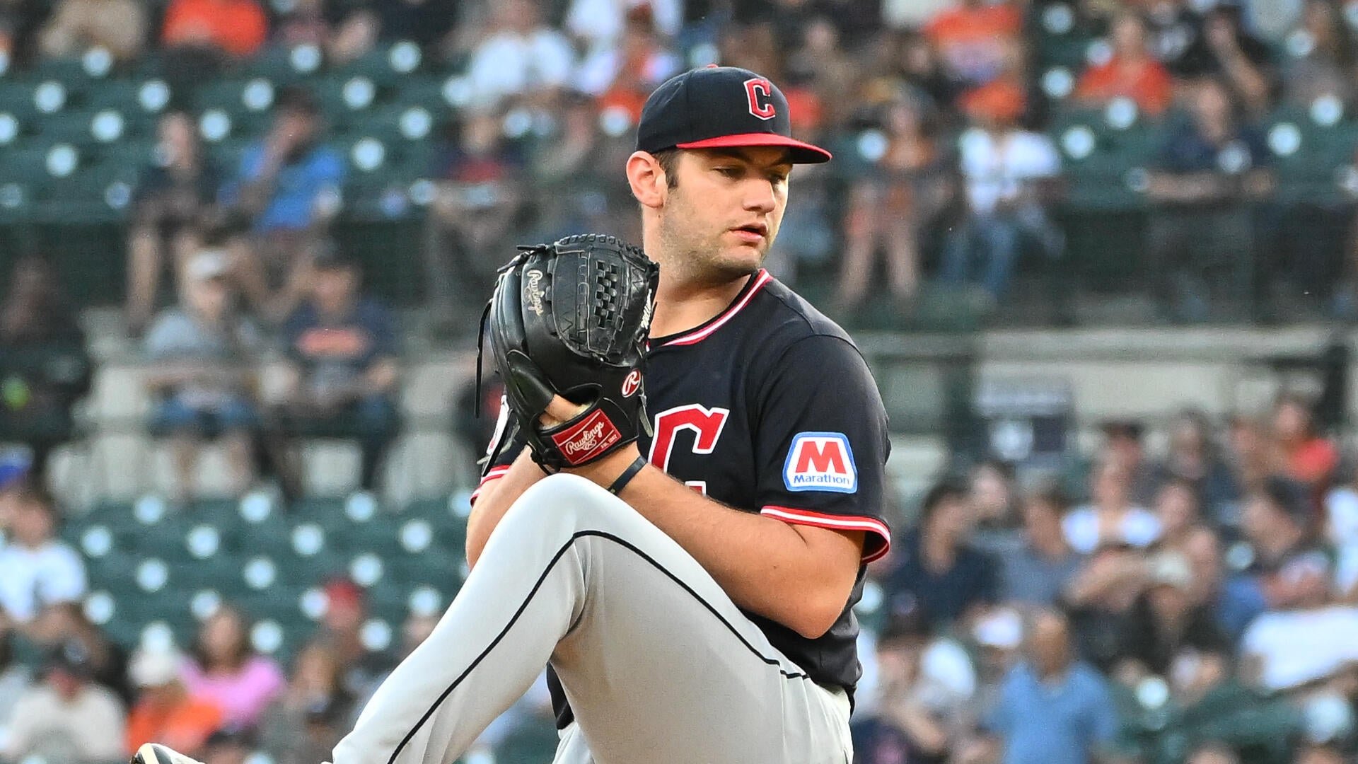 A baseball pitcher in a dark uniform with a "C" cap winds up with a glove on his left hand, set against a blurred crowd of stadium spectators.