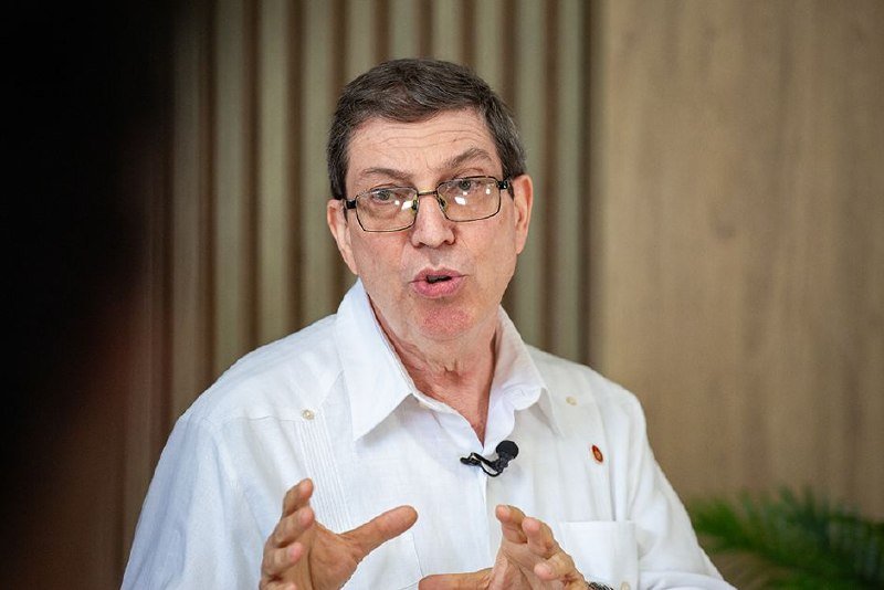 A man wearing glasses and a white shirt with a lapel microphone gestures with his hands while speaking in front of a wood-paneled background.