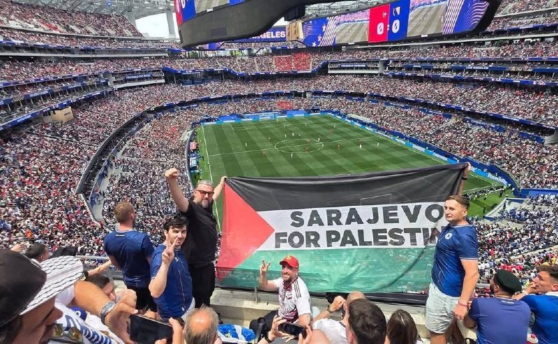Spectators in a packed stadium hold up a Palestinian flag and a banner reading "SARAJEVO FOR PALESTINE" during a soccer match.