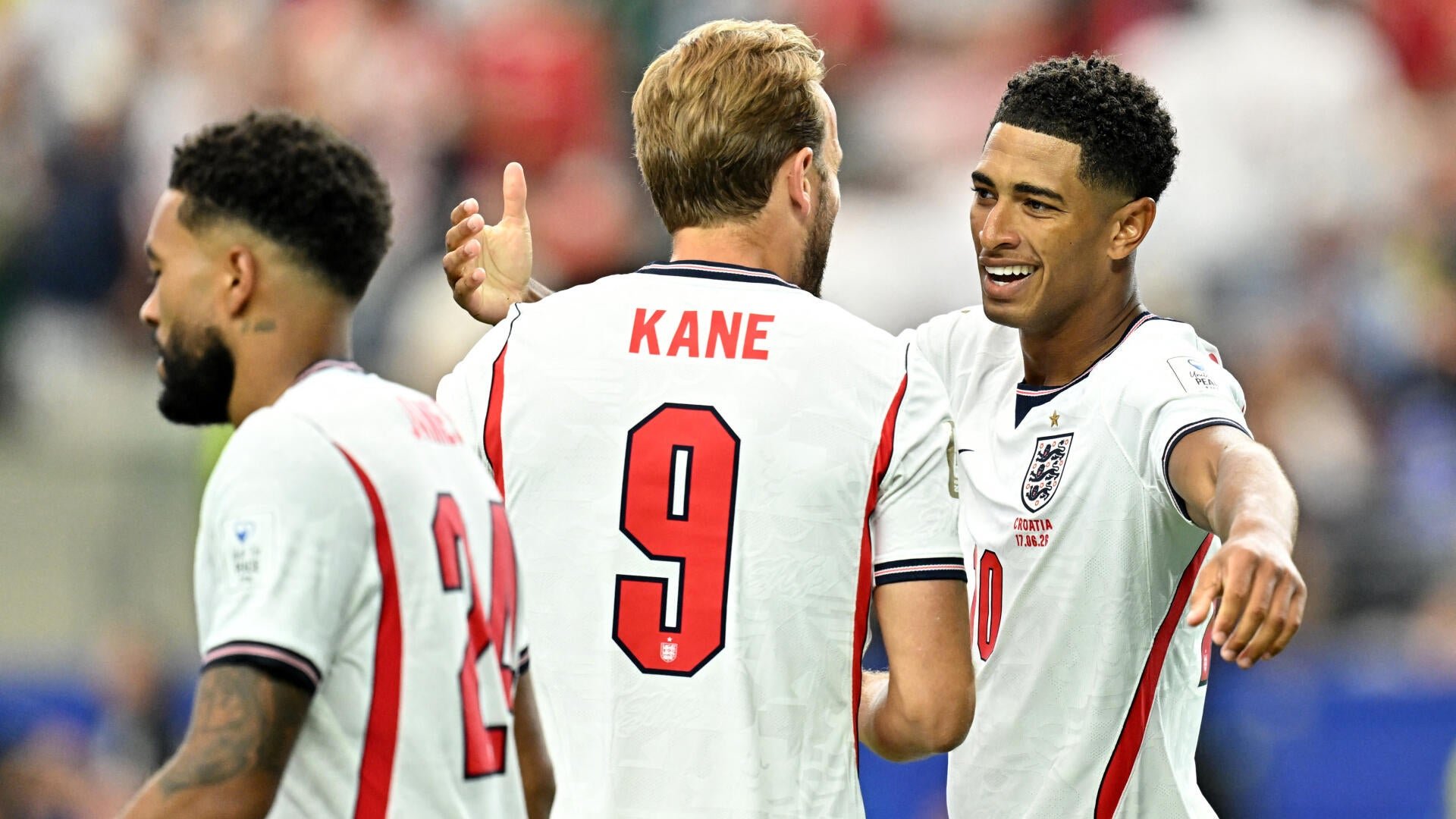 Three England soccer players in white jerseys celebrate together on a field, with one jersey displaying "KANE" and the number 9 on its back.