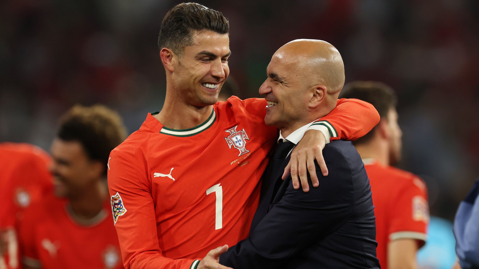 A male soccer player wearing a white US Soccer jersey stands in profile with a focused expression, a blurred figure and large digital screen visible in the background.