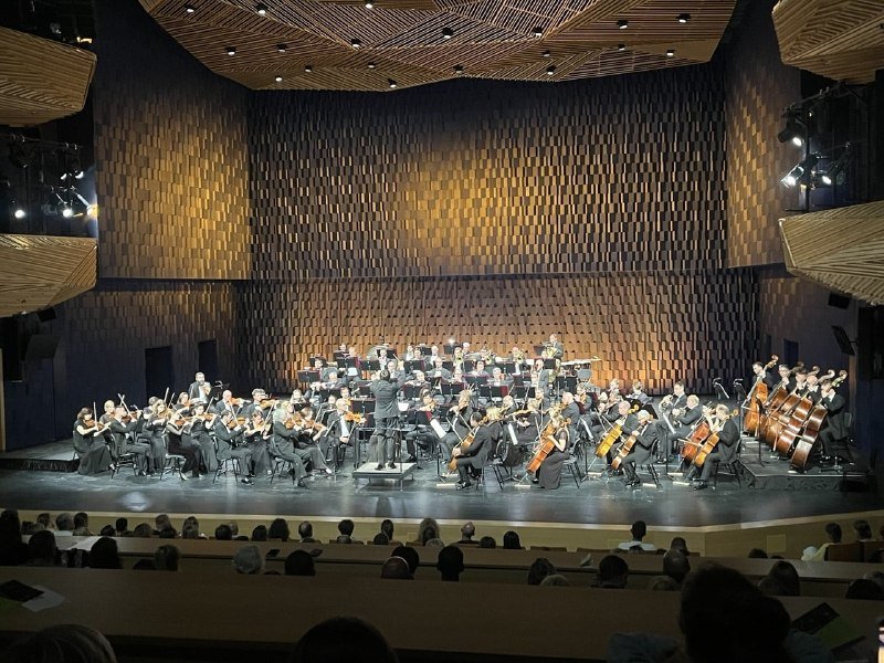 A symphony orchestra performs on a dimly lit stage in a modern concert hall with textured wooden walls, as a conductor leads seated musicians while an audience watches from below.