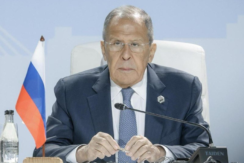 An older man in a dark suit and glasses sits at a table with a microphone and a small Russian flag, hands clasped together.