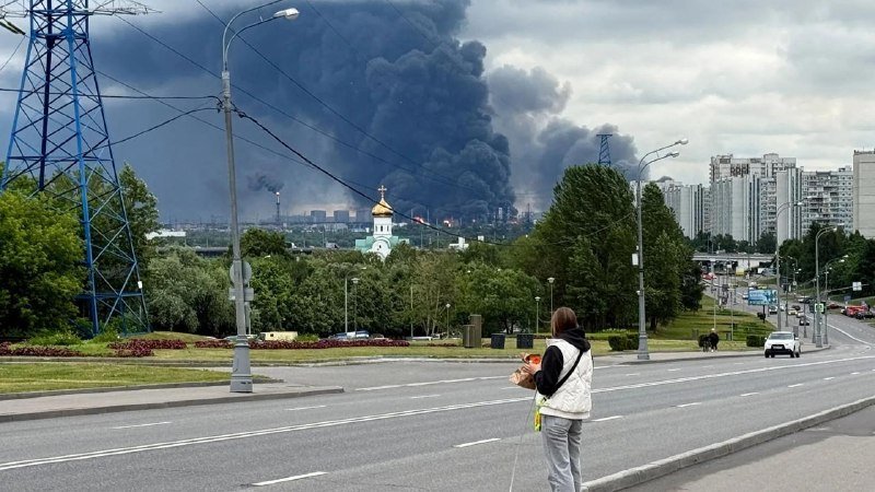 A hooded woman stands on a city roadside watching a massive plume of black smoke rising in the distance, with a small fire visible near the smoke's base.