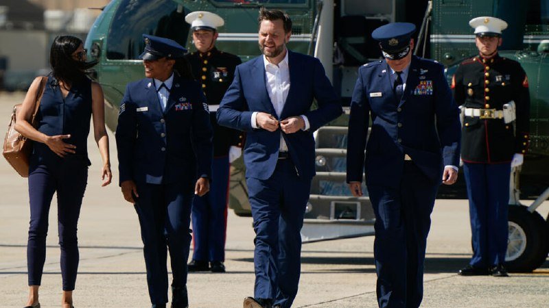 A man in a blue suit walks across a tarmac accompanied by two uniformed Air Force officers, with two Marines in dress uniform standing at attention near a green helicopter.