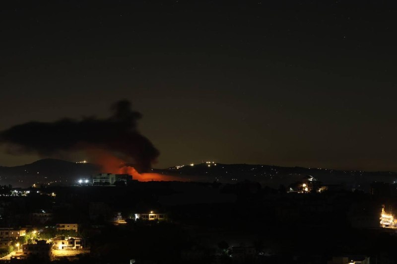 A plume of smoke rises above a distant town of low-rise buildings, viewed from behind a red-tiled rooftop under a clear blue sky.