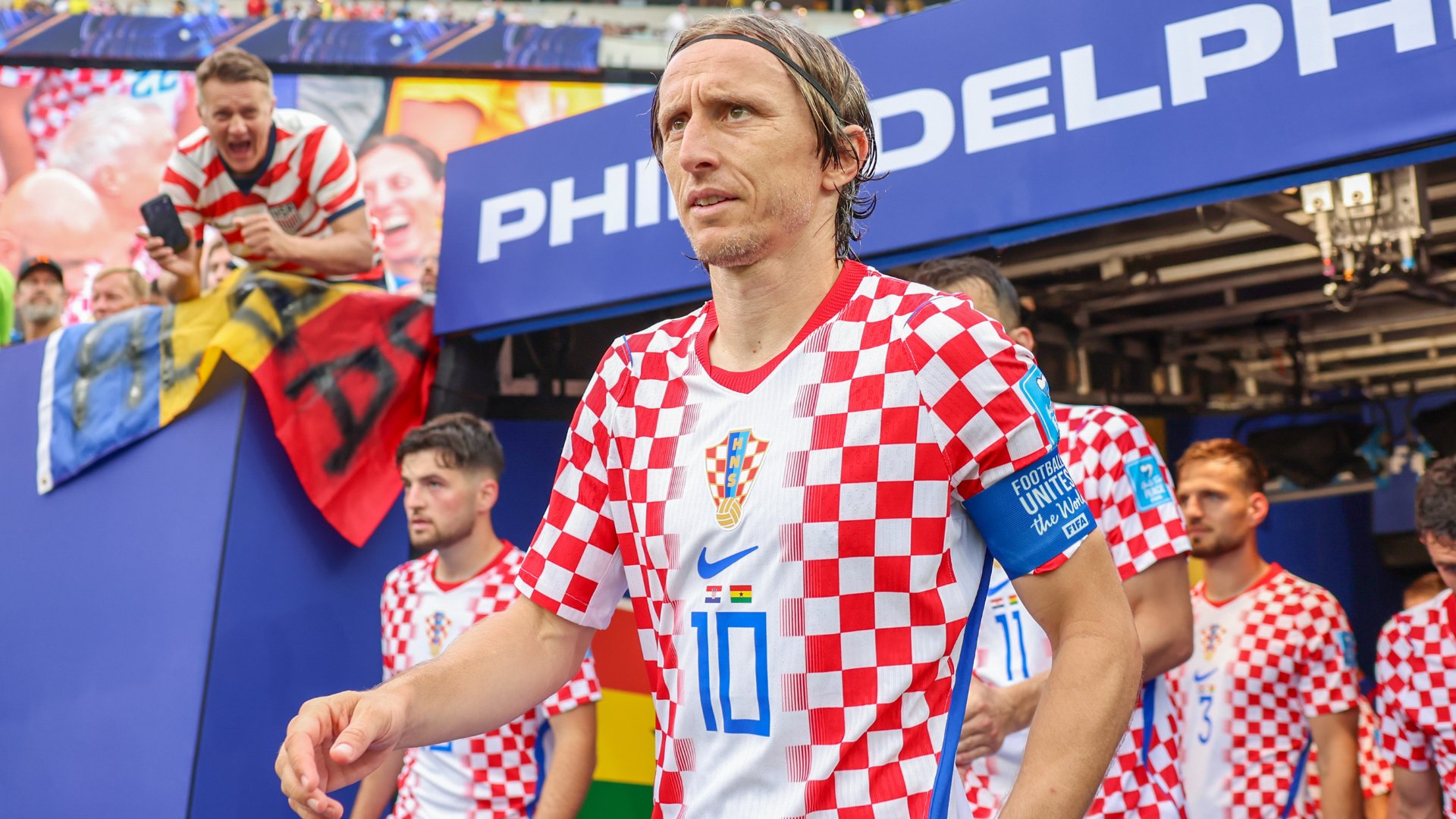 A long-haired soccer player wearing a red-and-white checkered jersey with the number 10 walks out of a stadium tunnel alongside teammates, as fans cheer behind a barrier.