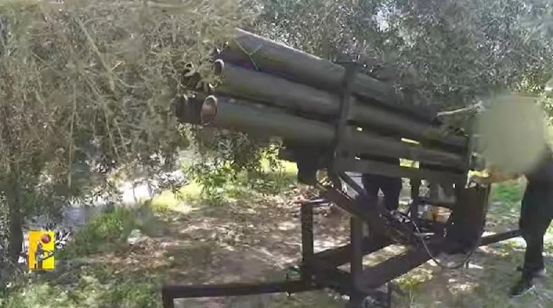 Multiple tube-style rocket launchers are mounted on a metal frame in a wooded outdoor setting, with partially visible figures standing nearby.