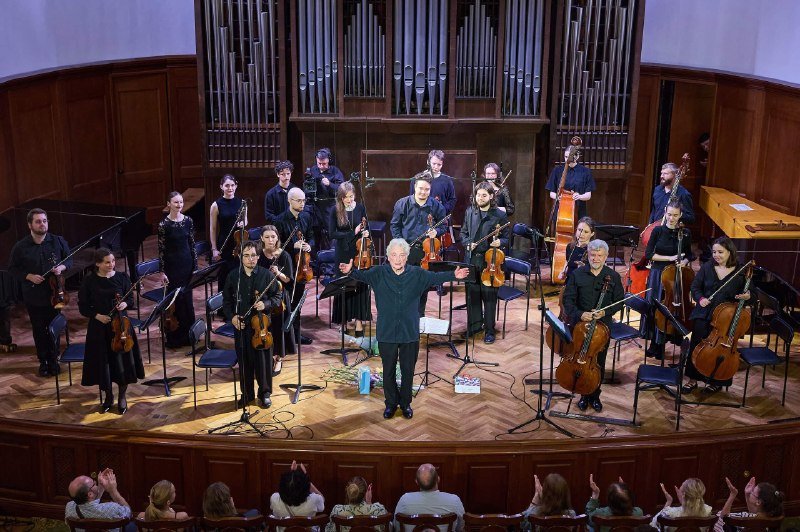 A conductor in dark attire stands center stage before a chamber orchestra of string and keyboard musicians inside a wood-paneled concert hall with large organ pipes.